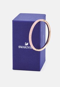 Ružovo-zlatý náramok s ružovými kryštálovými ozdobami, s hladkým povrchom a bezpečným zapínaním, vystavený na modrej Swarovski krabičke.