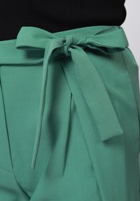 Zelene hlače iz tkanine z zapetim pasom na pasu, z gladko teksturo in pregibanim dizajnom, ki ujame sproščen slog.