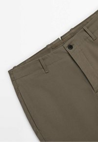Gros plan sur la ceinture d'un pantalon beige foncé avec passants pour ceinture, fermeture par bouton et braguette à glissière sur fond blanc.