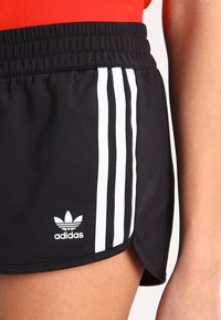Mustat urheilushortsit, joissa on teksturoitu kangas. Sivussa on kolme valkoista raitaa ja pieni valkoinen Adidas-logo helman lähellä.
