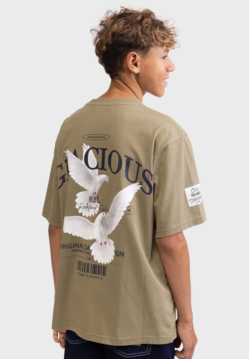 Olive green oversized t-shirt featuring white dove graphic, "GRACIOUS" text, barcode detail, and branded patch on the sleeve. Cotton material.