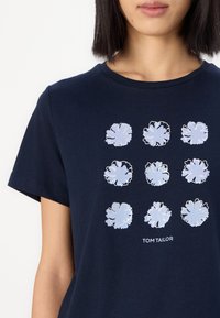 Mørkeblå t-skjorte med et blomstertrykkdesign i lyseblått, med ni blomsterformer ordnet i et rutenett. Merkenavnet "TOM TAILOR" under.