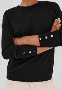 Maglione nero con polsini a coste e tre bottoni argentati decorativi, realizzato in un materiale morbido, con un semplice colletto a girocollo e vestibilità rilassata.