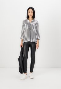 Gestreepte, lichte button-up shirt in grijs en wit, gecombineerd met zwarte, glanzende leggings. Model houdt een zwarte totebag vast en draagt witte sneakers.