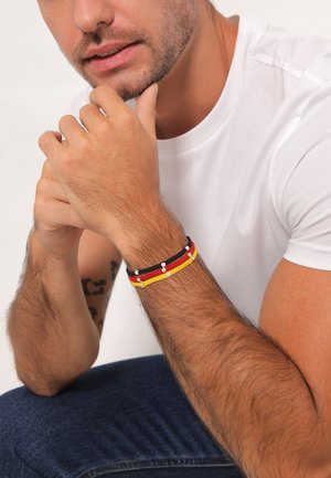 Farverig vævet armbånd med sorte, røde og gule tråde, pyntet med små hvide perler; båret på et håndled med lys hudtone.