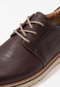 Chaussure en cuir marron avec une finition texturée, dotée de lacets beiges, d'une semelle claire contrastante et de coutures visibles le long des bords.