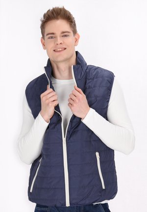 Laikavaimenta vests sinis ar augstu apkakli, ar rāvējslēdzēju un divām sānu kabatām. Modelis valkā baltu garām piedurknēm kreklu zem tā.