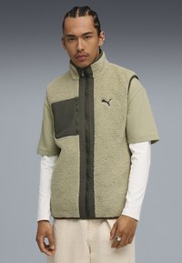Gilet in felpa verde con zip frontale, pannellatura in verde scuro e logo. Indossato sopra una camicia in tessuto verde chiaro e una maglietta a maniche lunghe bianca.