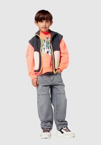 Canotta grigia senza maniche sopra una felpa arancione con stampa grafica, pantaloni baggy grigi e scarpe bianche con accenti rossi e neri.