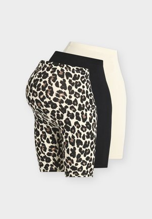 Donna in piedi che indossa un top nero senza maniche, pantaloncini biker con stampa leopardata, scarpe slingback nere con borchie e tiene una grande borsa tote nera.