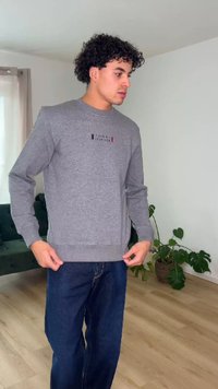 Mladý muž s kučeravými vlasmi, ktorý nosí sivú mikinu a modré džínsy, stojí v miestnosti s drevenou podlahou a rastlinami.