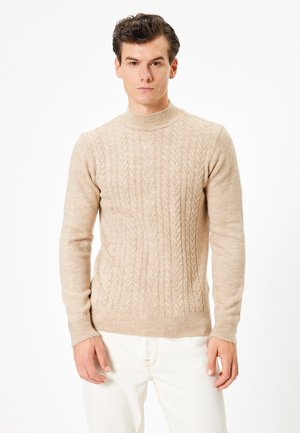 Suéter beige de punto con cuello alto, patrón texturizado, puños y dobladillo acanalados, confeccionado en material suave, llevado sobre unos jeans de color claro.