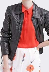 Schwarze Lederjacke mit taillierter Passform, ausgestattet mit Reißverschlusstaschen, scharfem Kragen und strukturiertem Finish, getragen über einer leuchtend roten Bluse.