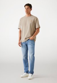 Lys beige bomulds T-shirt med et brystlogo, parret med lyseblå denimjeans. Modellen har hvide sneakers på og står foran en neutral baggrund.