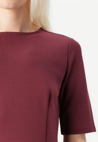 Top scurt cu maneci din burgund, fabricat dintr-un material neted, având un decolteu rotund cu detalii de cusătură și o formă ajustată. Nu sunt vizibile modele sau accente.