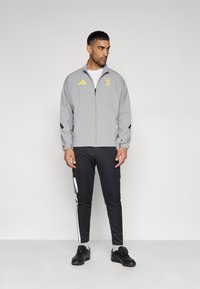 Γκρι αθλητικό μπουφάν με κίτρινο λογότυπο και μαύρες λεπτομέρειες, σε συνδυασμό με μαύρο παντελόνι με λευκές ρίγες. Φορώντας μαύρα παπούτσια.