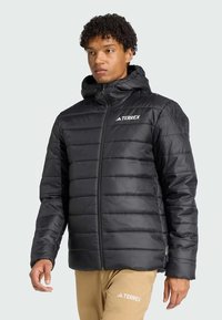 Čierna prešívaná vetrovka s kapucňou, predným zipsom, vzorom prešívania a logom Terrex na ľavej strane hrude; hladký, lesklý materiál.