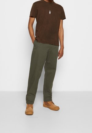 Braunfarbenes T-Shirt mit kurzen Ärmeln und einem kleinen weißen Grafikmotiv, kombiniert mit olivgrünen Hosen und braunen Stiefeln mit strukturiertem Finish.