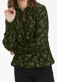 Blouse verte à motifs géométriques, encolure à volants et fermeture à nouer. Comprend des manches longues avec poignets froncés et tissu texturé.