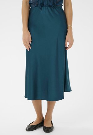 Femme portant une jupe midi bleu sarcelle et des ballerines noires, debout avec les mains détendues le long du corps sur fond blanc.