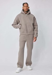 Personne debout portant un sweat à capuche taupe avec fermeture éclair et un pantalon assorti, ainsi que des baskets blanches, sur un fond clair uni.