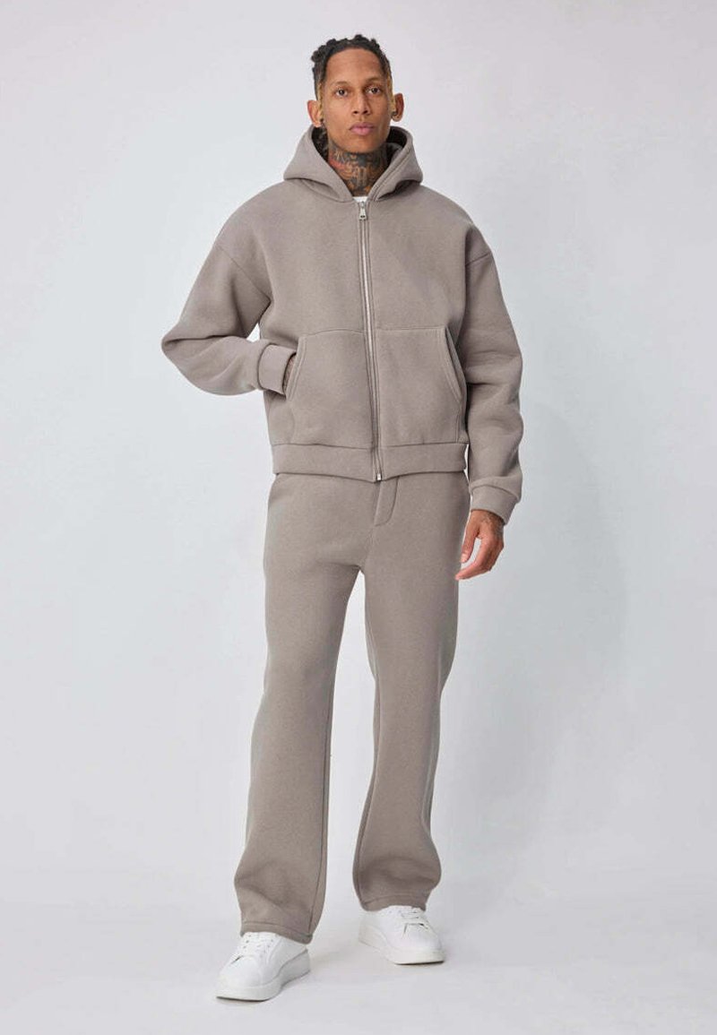 Personne debout portant un sweat à capuche taupe avec fermeture éclair et un pantalon assorti, ainsi que des baskets blanches, sur un fond clair uni.
