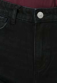 Svarte denimjeans med en sølvfarget metallknapp, beltehemper og lommer med en lett strukturert overflate. Nærbilde av livet.