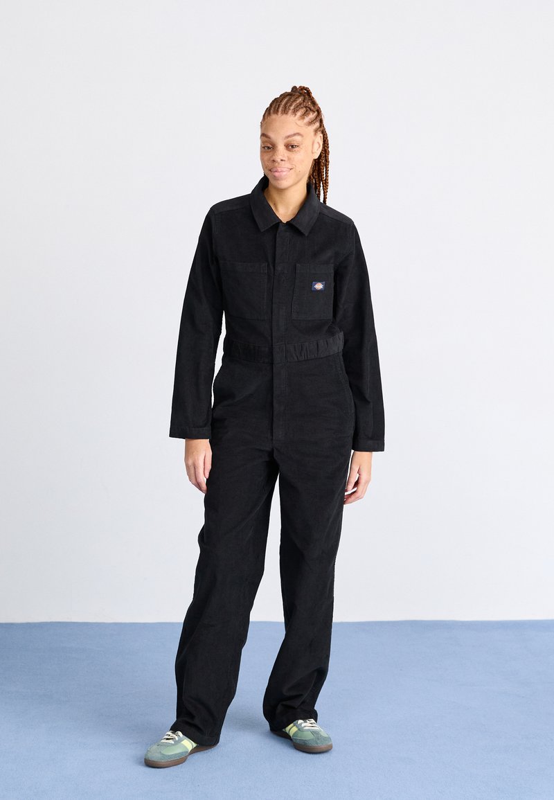 Salopette en velours côtelé noir avec des manches longues, un col, des poches latérales et une taille cintrée, associée à des baskets vertes et grises.