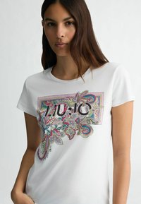 T-shirt bianco a maniche corte con un vivace motivo paisley e una grafica audace "10" al centro. Tessuto di cotone morbido e liscio.