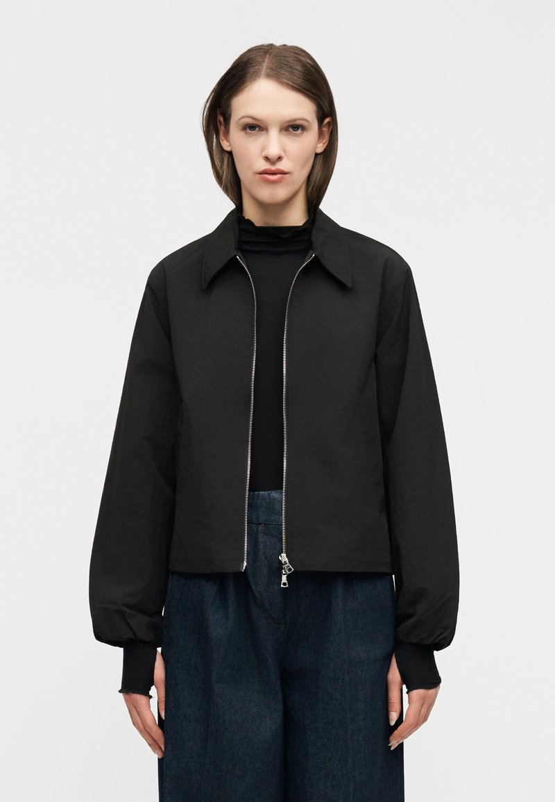 Γυναίκα που φοράει μαύρο μπουφάν μ ε φερμουάρ πάνω από μαύρο τοπ και φαρδιά σκούρα μπλε παντελόνια, στέκεται μπροστά σε ένα απλό φωτεινό φόντο.