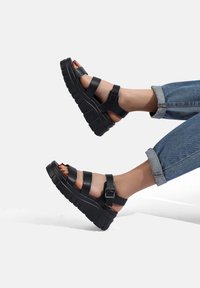 Sandali platform neri con cinturini spessi e fibbia. Presentano un design open toe e una suola texturizzata. Abbinati a jeans di denim con risvolto.