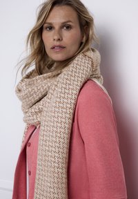 Beżowy szal o fakturowanym, tkanym wzorze z jasnobrązowymi akcentami, zarzucony na szyję i zestawiony z koralowym sweterkiem.