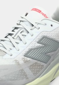 Gray athletic shoe with a textured mesh upper, prominent gray “N” logo, lace-up closure, and a white and gray sole. Red branding on the tongue.