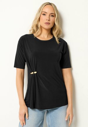 Femme blonde portant un haut noir à manches courtes avec un embellissement feuille d'or à la taille, associée à un jean bleu clair, debout contre un mur blanc.
