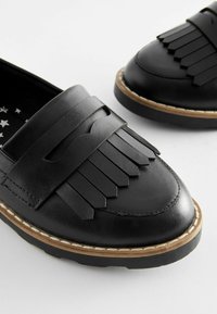 Schwarze Lederlounge-Schuhe mit Fransenverzierungen, runder Zehenpartie, weißem gesticktem Rand und einer strukturierten Gummisohle. Das Innere ist mit einem Sternmuster versehen.