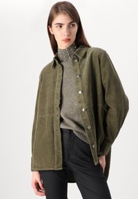 Casaco de camisa de corduroy verde-azeitona com botões metálicos, apresentando um corte descontraído e uma superfície texturizada, sobre uma blusa de malha cinza.