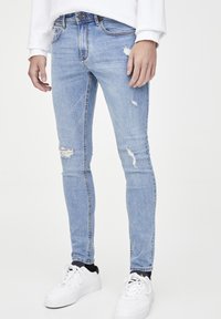 Jeans skinny de color azul claro con detalles desgastados y un ligero desvanecimiento, hechos de denim. Combinados con zapatillas blancas y un sudadera blanca.