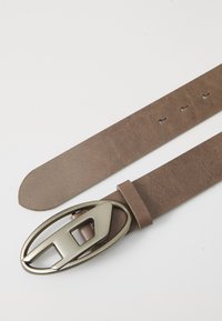 Cinturón de cuero marrón con una textura suave, que presenta una hebilla de metal ovalada con un diseño de corte geométrico. Ajustable con agujeros perforados.