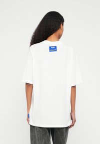 Άσπρο βαμβακερό T-shirt με χαλαρή εφαρμογή, με μ πλε ετικέτα μάρκας στην πλάτη και διακριτική πλαϊνή ετικέτα. Σκούρο γκρι παντελόνι με υφή.