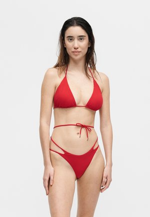 Kvinne med langt mørkt hår iført en rød todelt bikini med halterneck og utskårne høye deler, stående mot en ensfarget lys bakgrunn.