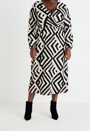 Kvinde iført en langærmet, sort-hvid midi kjole med geometrisk mønster og sorte ankelstøvler, stående mod en hvid baggrund.