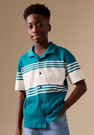 Adolescent debout, main dans la poche, portant une chemise à manches courtes bleu sarcelle et blanc à rayures et un jean bleu clair.