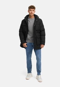 Giacca nera isolata con un design strutturato, due tasche frontali e cappuccio, indossata sopra un maglione grigio e jeans azzurri chiari.