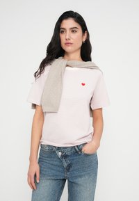 Ροζ ριγέ μπλουζάκι με κόκκινη καρδιά κεντημένη στο αριστερό στήθος, συνδυασμένο με μπεζ πουλόβερ που είναι περασμένο πάνω από τους ώμους. Φοριέται με μπλε τζιν.