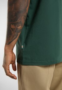 Zelené bavlněné tričko se rovnými střihy, krátkými rukávy a božní nášivkou. Zobrazuje viditelné tetování na paži a béžové kalhoty.