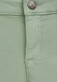 Svetlo zelena denimska tkanina z gladko teksturo, ki ima srebrn gumb in šivane šive v bližini območja žepa.