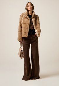 Chaqueta de faux fur en beige con rayas horizontales, combinada con pantalones de pierna ancha en marrón oscuro. La modelo sostiene un bolso marrón claro.