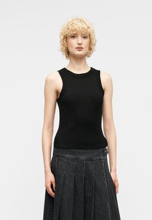 Άτομο με ξανθά, σγουρά μαλλιά, ντυμένο με μαύρη, αμάνικη μπλούζα και σκούρα φούστα με πτυχώσεις και ψηλή μέση, μπροστά σε απλό φόντο.