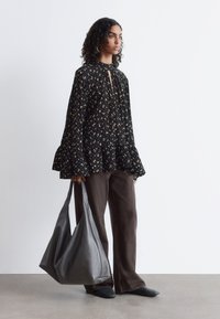 Schwarze Bluse mit Blumenmuster und Glockenärmeln, kombiniert mit braunen weit geschnittenen Hosen. Große graue Ledertasche in der Hand, schwarze Slipper getragen.