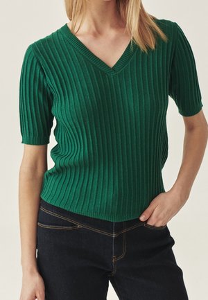 Woman wearing a fitted green ribbed short-sleeve V-neck sweater with dark blue jeans, one hand resting in pocket.